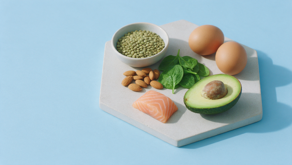 Lentilles, amandes, œufs, épinards, avocat et saumon posés sur un plateau de pierre hexagonal, sur fond bleu ciel