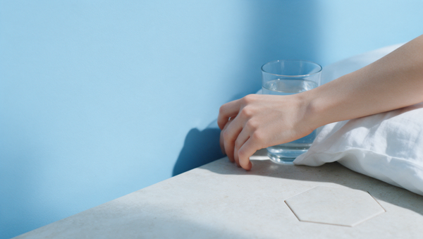 Main posée près d'un verre d'eau sur une table de chevet claire au réveil, oreiller blanc et mur bleu ciel